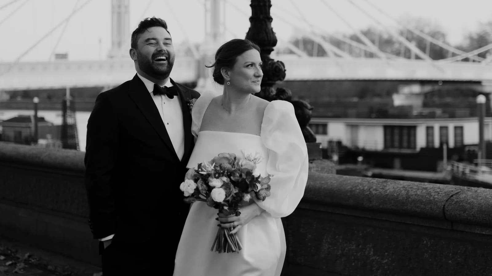 Bride and groom stand next to bridge on Thames during their winter wedding in London