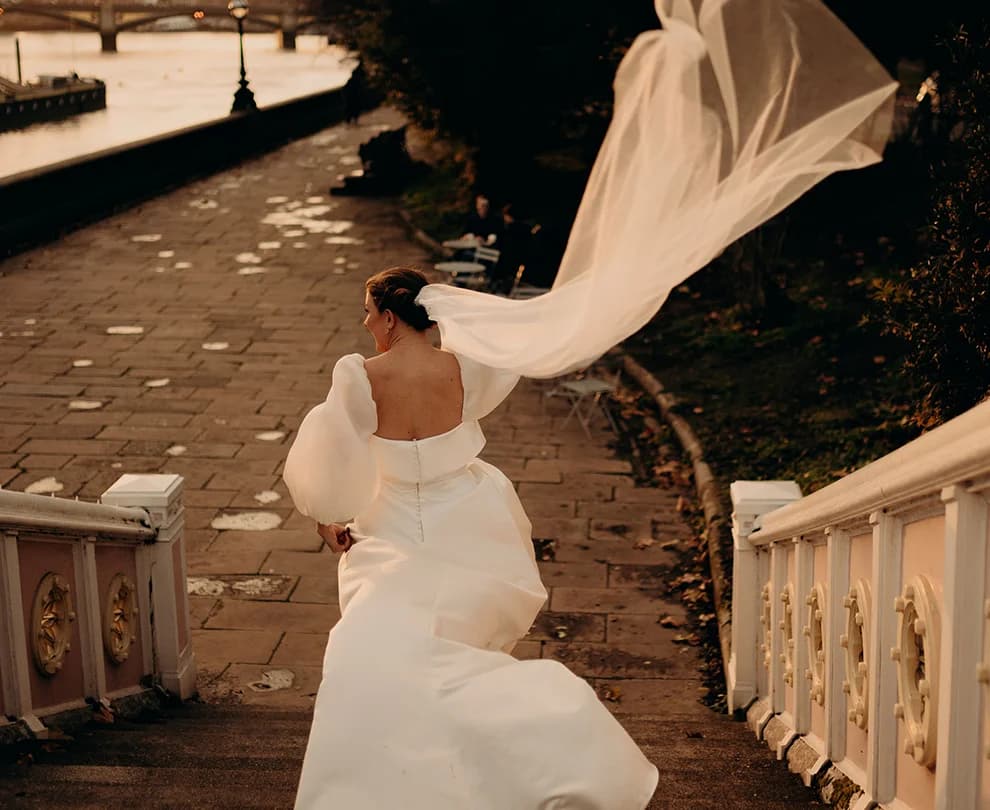 Bride walks down the stairs next to Thames while her veil is blown in the air by wind