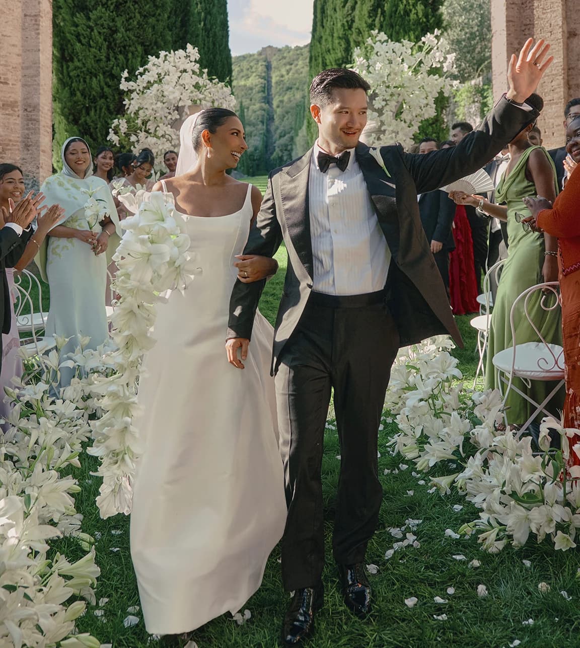 Outdoor wedding recessional at an Italian villa, featuring a groom in a classic black tuxedo and bow tie walking with the bride through a cypress-lined aisle.