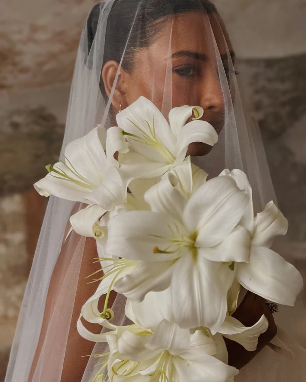Close-up bridal portrait featuring a bouquet of large white lilies, softly framed by a sheer veil against a warm, painterly backdrop.