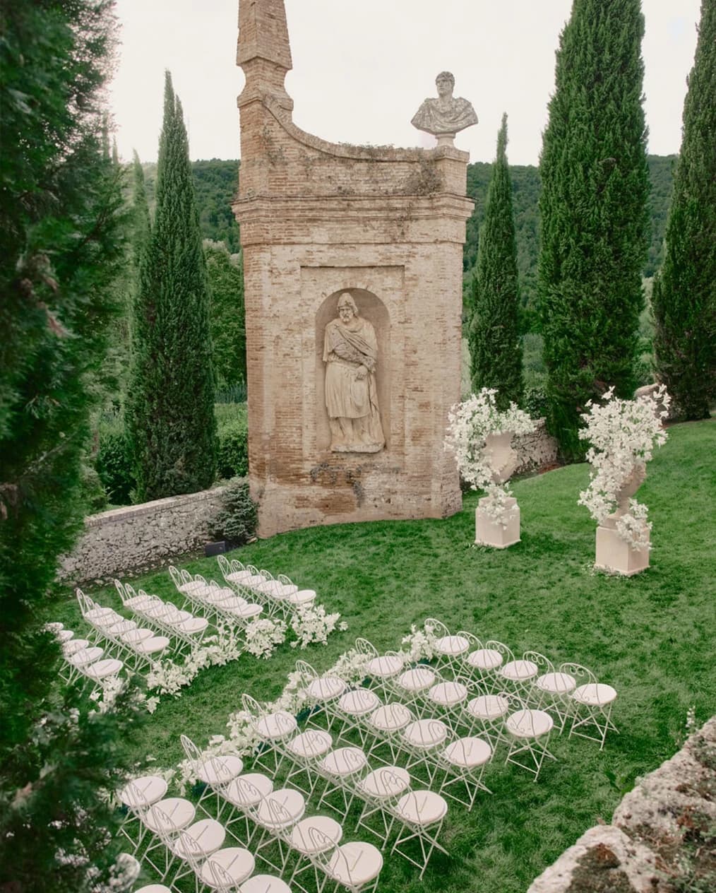 Wedding ceremony setup at an Italian villa, with rows of white chairs and floral arrangements set against historic stone architecture and cypress trees.