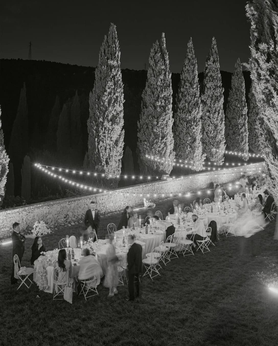 Evening wedding reception set beneath string lights, with guests dining at long tables surrounded by cypress trees in a romantic outdoor setting.