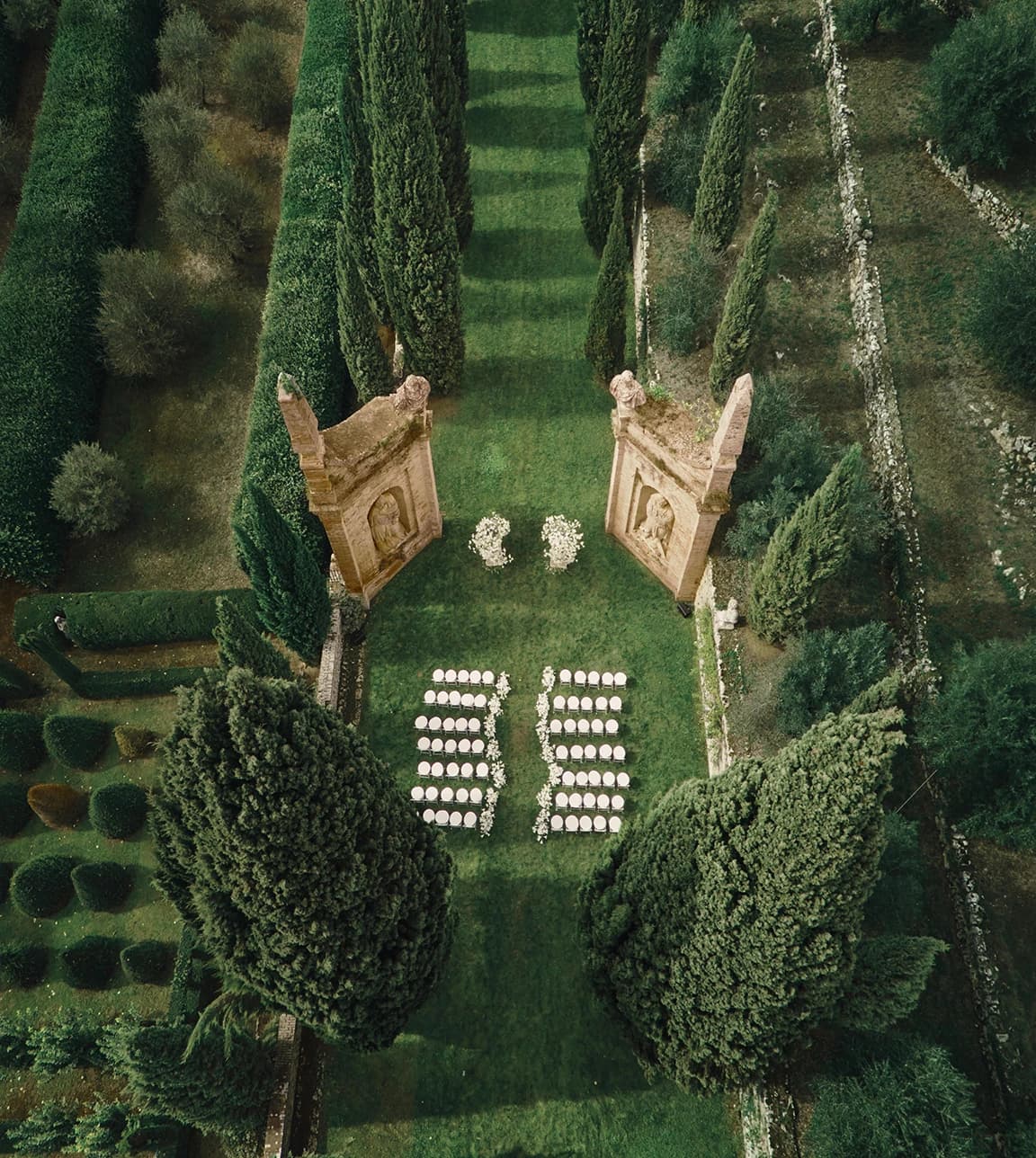Aerial view of an Italian villa garden ceremony setup, with a cypress-lined aisle, symmetrical seating, and floral arrangements framed by historic stone arches.