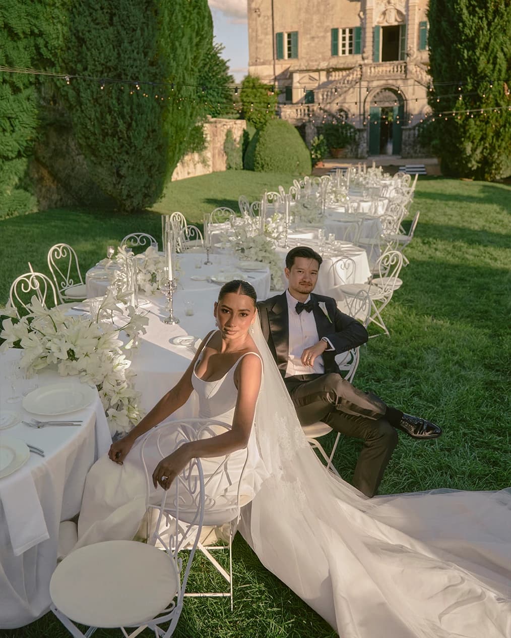 Outdoor wedding reception at Villa Cetinale, with a long, elegantly styled table adorned with white florals, as the bride and groom sit together in the sunlit garden