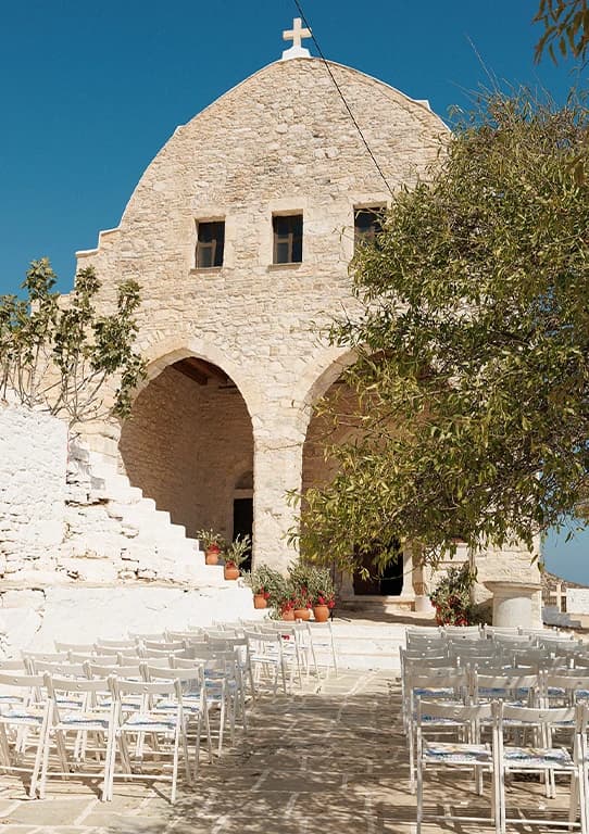 Outdoor wedding ceremony setup with white chairs in front of a traditional stone chapel on a Greek island.