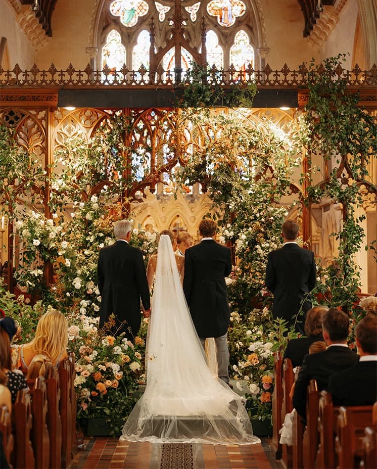 Bride and groom exchanging vows during a luxury church wedding ceremony surrounded by abundant floral installations and family members.