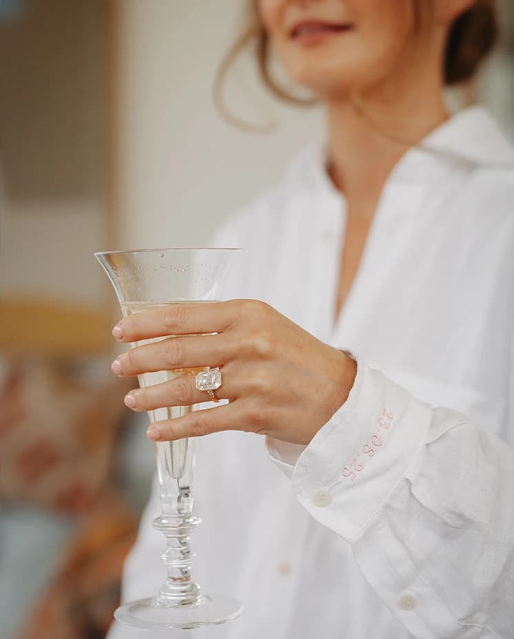 Bride holding a champagne glass during wedding preparations, showing her engagement ring and an embroidered wedding date on her shirt cuff.