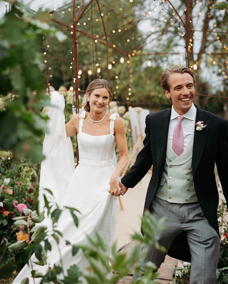 Bride and groom walking hand in hand through a flower-filled garden walkway illuminated by twinkling lights during a luxury countryside wedding reception.