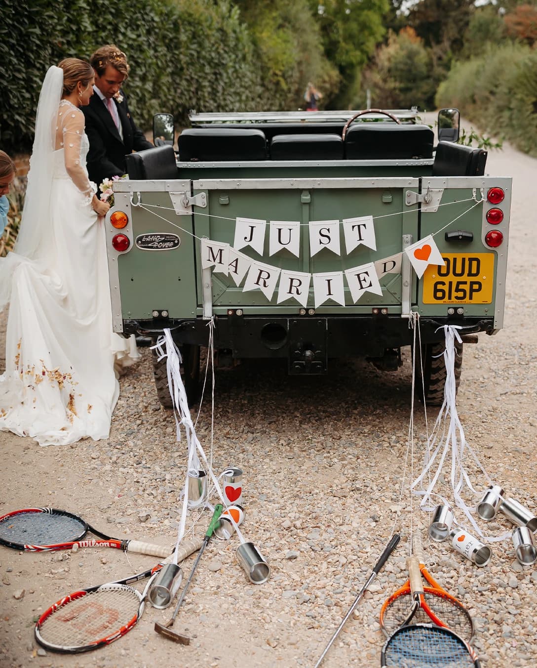 Bride and groom beside a vintage Land Rover decorated with a 'Just Married' sign, trailing tin cans and tennis rackets after their countryside wedding ceremony.