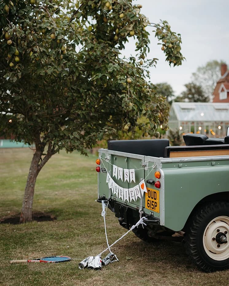 Vintage Land Rover wedding car decorated with a 'Just Married' sign, trailing tin cans and a tennis racket parked beneath a pear tree at a countryside wedding venue.
