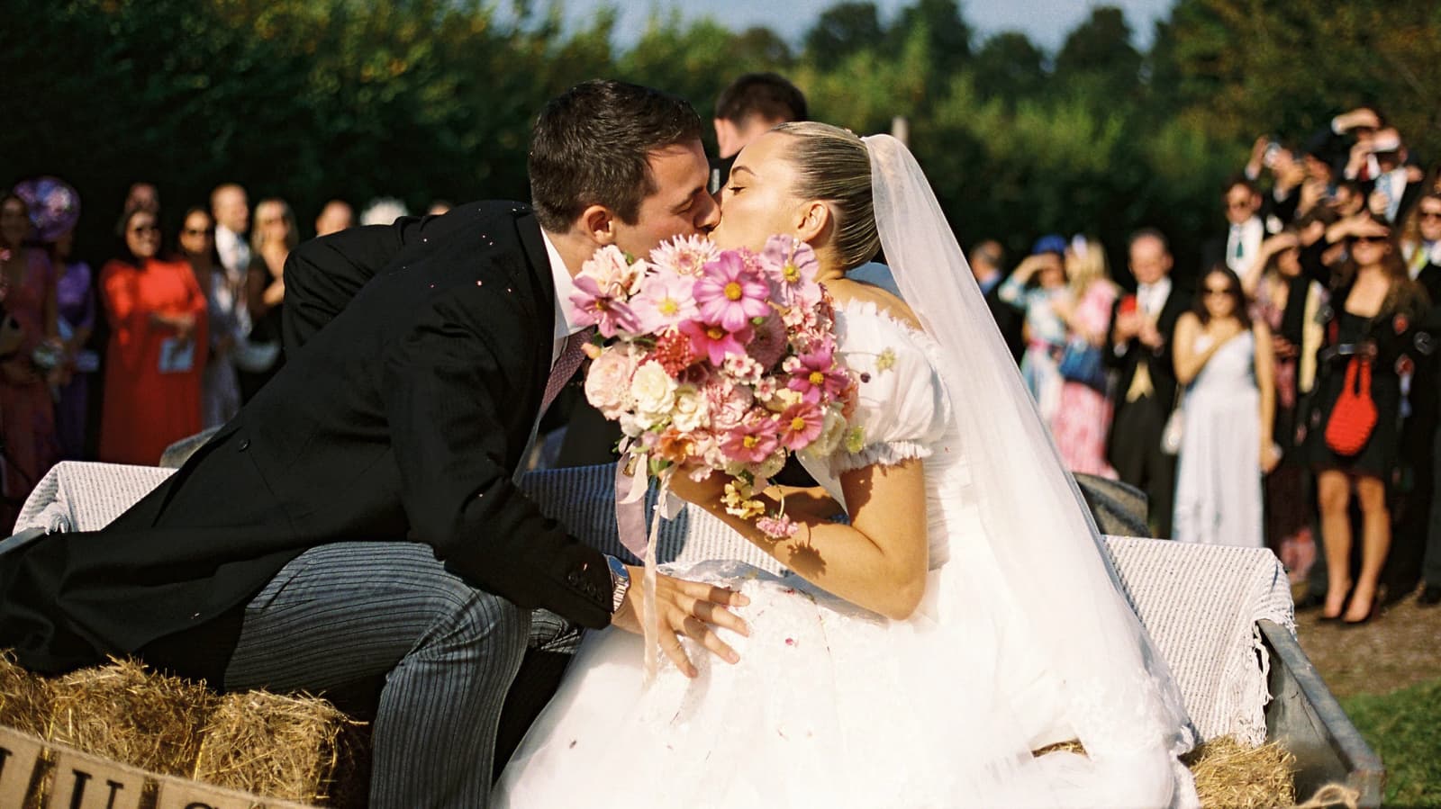 Bride and groom sharing a joyful kiss in a flower-filled wedding tractor after their English countryside wedding ceremony, surrounded by family and friends.