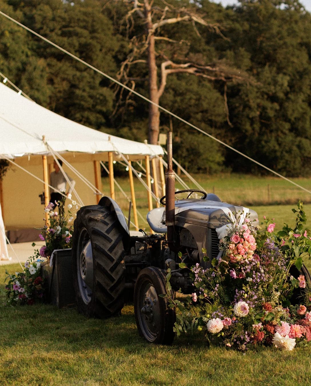 Vintage tractor decorated with colourful floral arrangements beside a marquee at an English countryside wedding reception. 