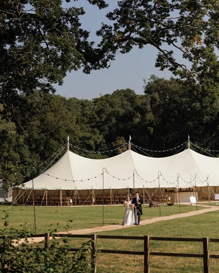 Bride and groom walking towards a sailcloth marquee at their English countryside wedding reception, surrounded by scenic woodland and open fields.