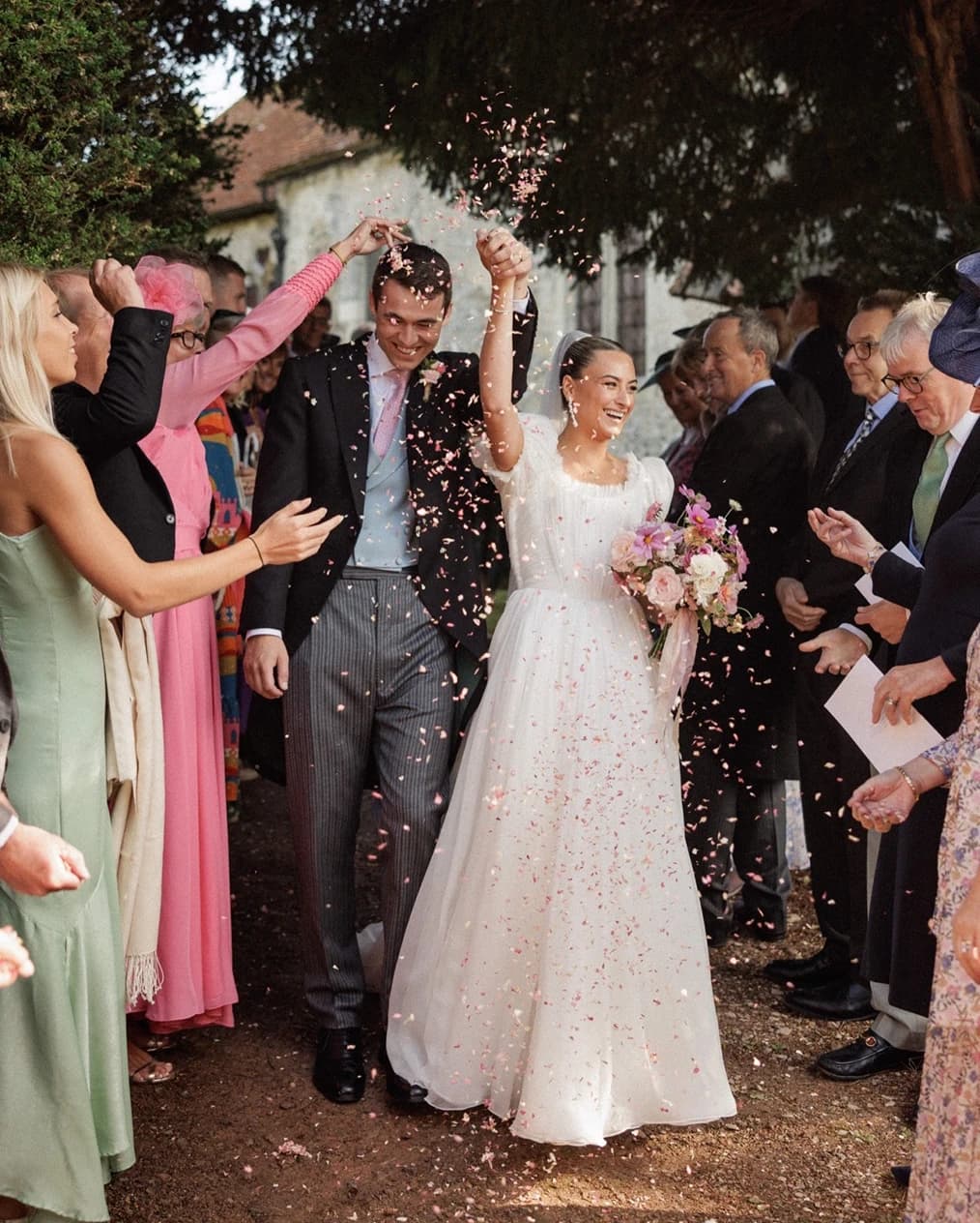 Bride and groom celebrating with a joyful confetti exit outside a historic English church, surrounded by family and friends after their wedding ceremonyt.