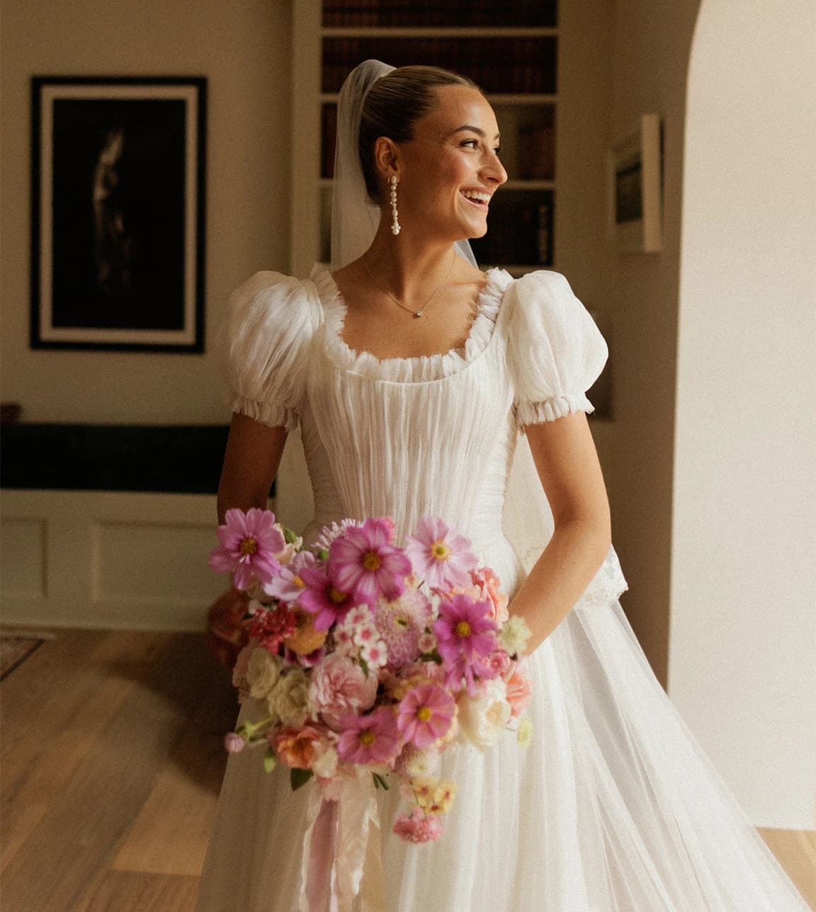 Bride in a romantic puff-sleeve wedding dress holding a vibrant pink bridal bouquet, smiling by a window before her English countryside wedding ceremony.