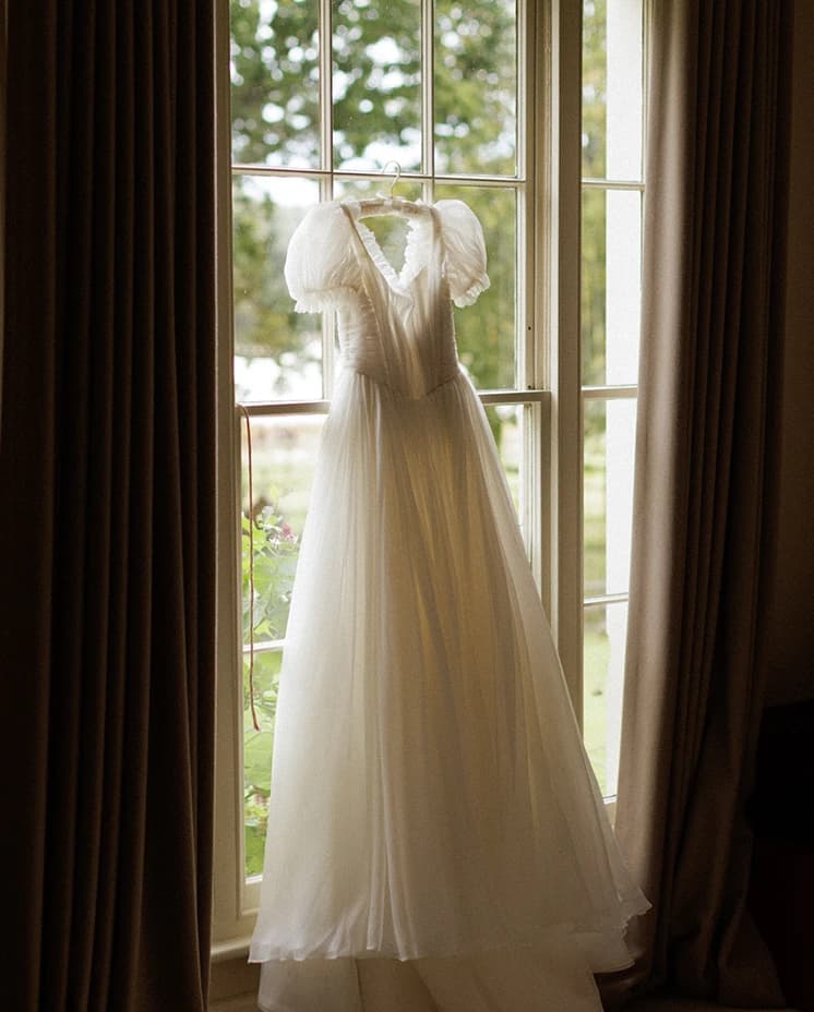 Elegant Emma Beaumont Design puff-sleeve wedding dress hanging in front of a window, beautifully displayed before an English countryside wedding ceremony.
