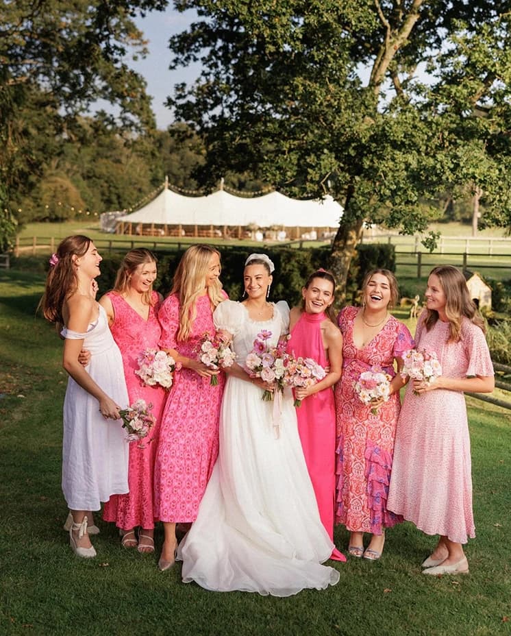 Bride and bridesmaids holding pink and blush bouquets in front of a sailcloth marquee, celebrating an elegant English countryside wedding with joyful group portraits.
