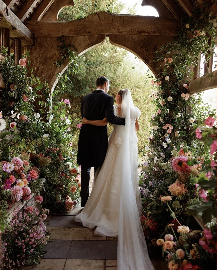 Bride and groom embracing beneath a flower-filled historic church lychgate, surrounded by romantic floral arrangements after their English countryside wedding ceremony.