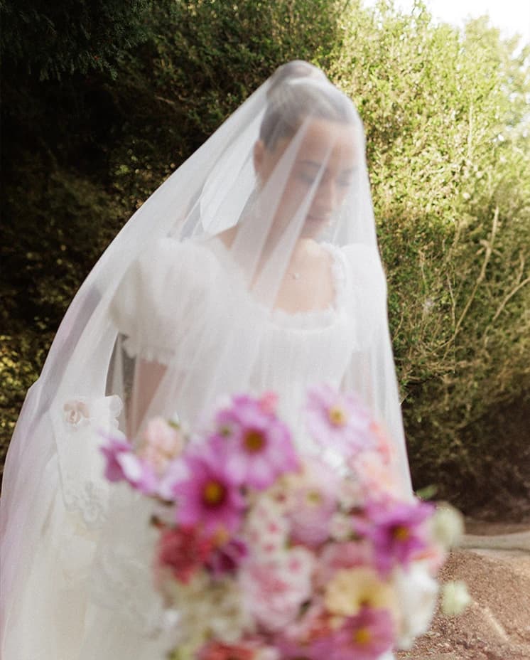 Bride wearing a delicate lace-edged veil and romantic puff-sleeve wedding dress, holding a pink bridal bouquet during an elegant English countryside wedding.