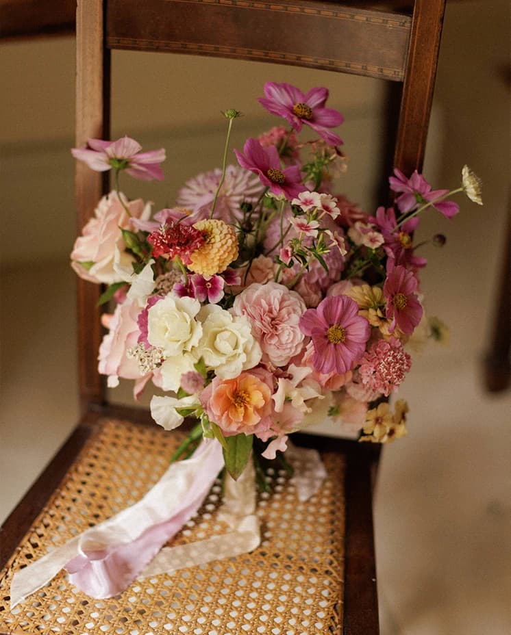 Romantic pink and blush bridal bouquet with seasonal flowers resting on a rustic wooden chair, styled for an elegant English countryside wedding.