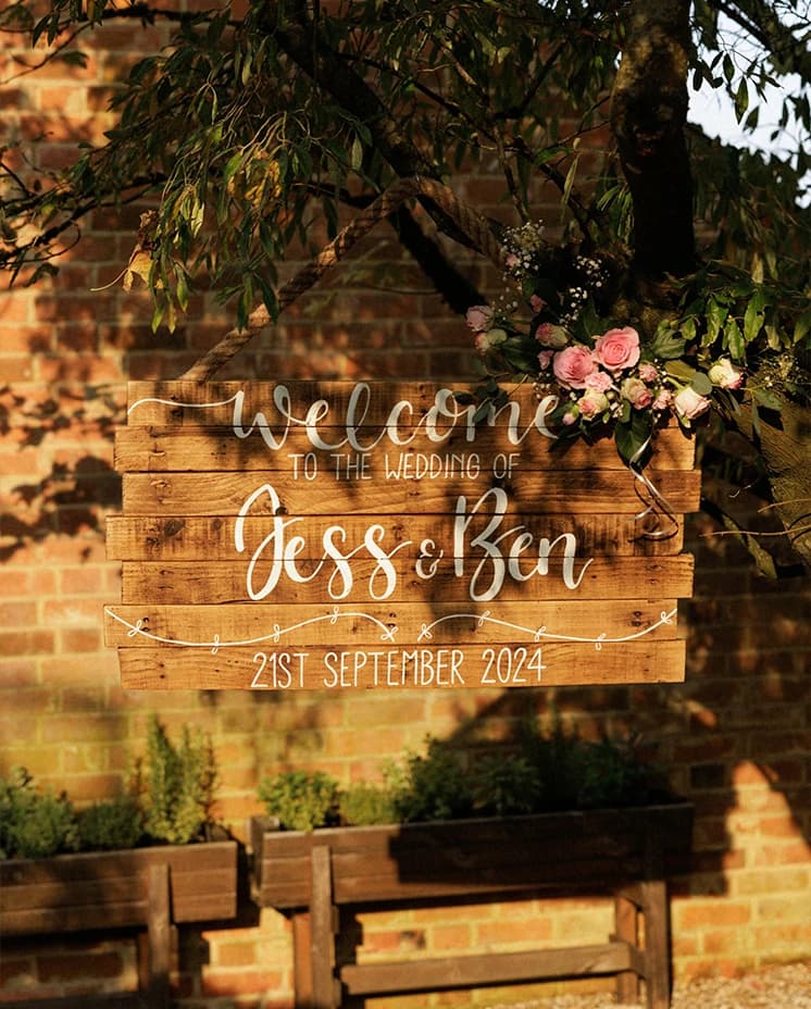 Rustic wooden wedding welcome sign decorated with pink flowers, welcoming guests to an elegant English countryside wedding venue.
