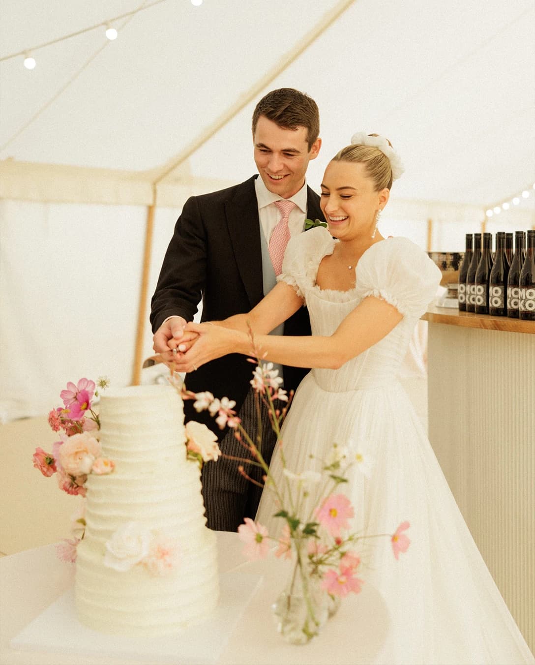 Bride and groom cutting their wedding cake inside a sailcloth marquee, celebrating their English countryside wedding with elegant floral styling.