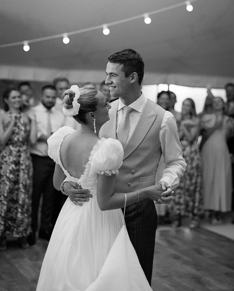 Bride and groom sharing their first dance beneath festoon lights in a sailcloth marquee, celebrating their English countryside wedding reception in an elegant black and white portrait.
