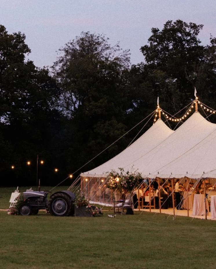 Sailcloth wedding marquee illuminated with festoon lights at dusk, with a vintage tractor creating a romantic English countryside wedding reception setting.