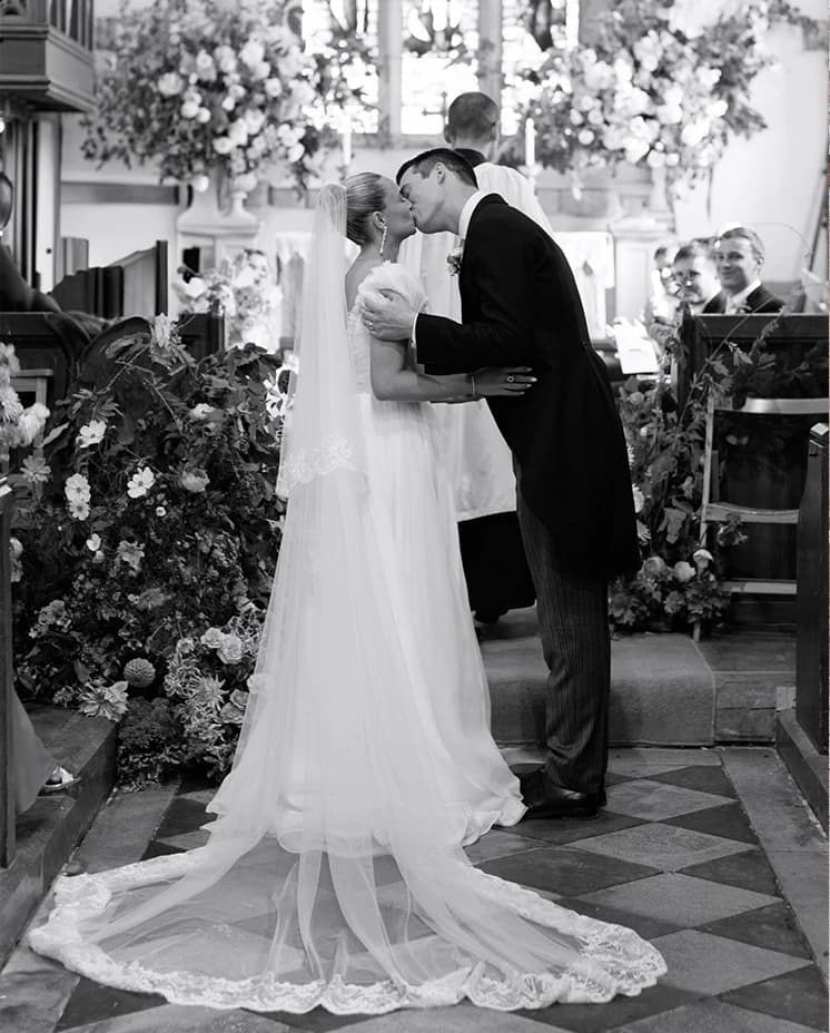 Bride and groom sharing their first kiss at the altar in a historic English church, surrounded by abundant floral arrangements during a romantic countryside wedding ceremony.