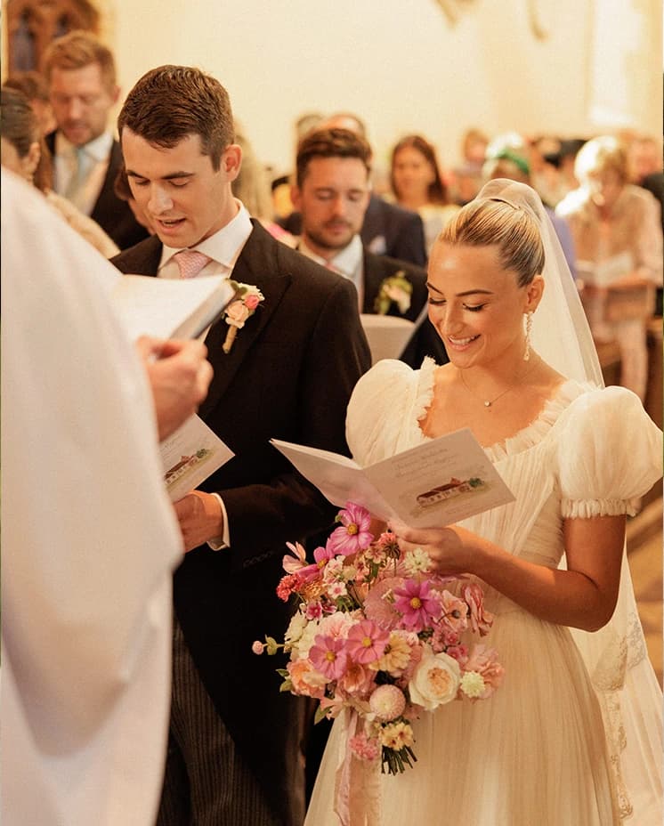 Bride and groom reading their vows during a romantic church wedding ceremony, with the bride holding a pink and blush bouquet in a historic English countryside church.