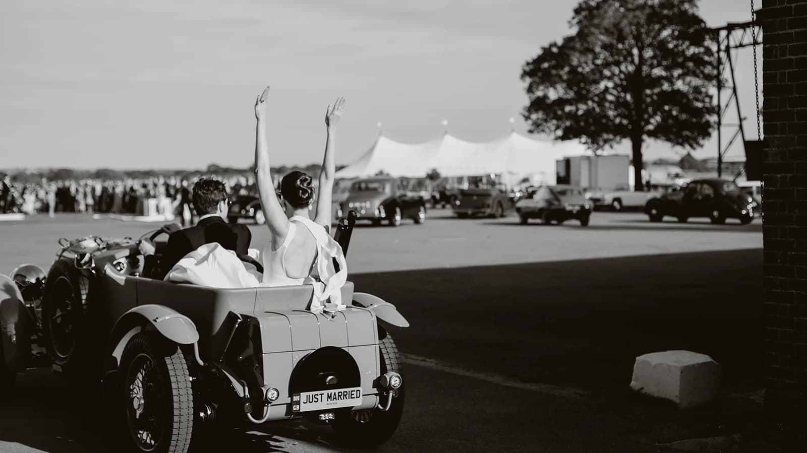 Bride raising her arms in celebration from the back of a vintage car on the airfield