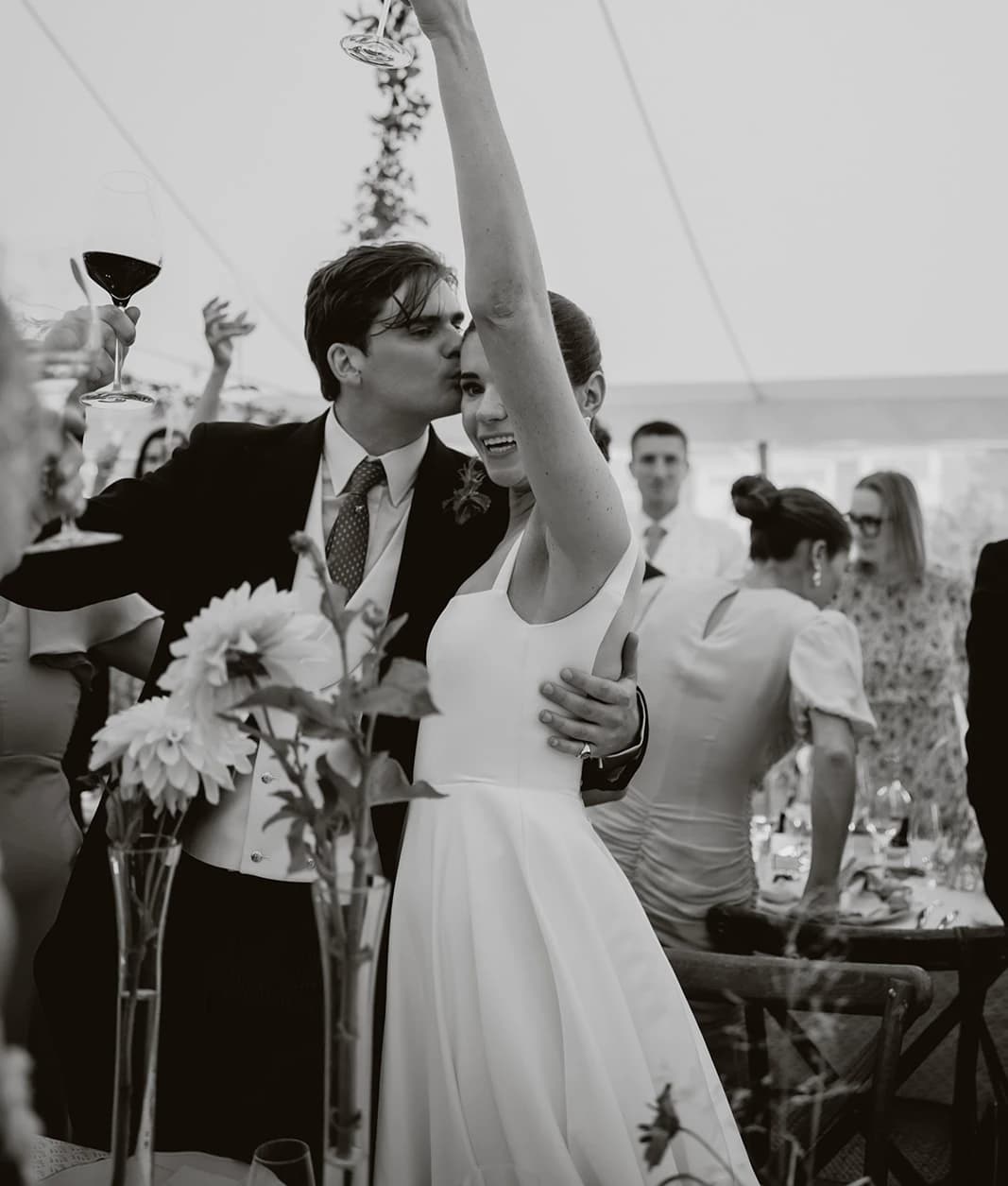 Black and white photograph of the bride raising her glass as the groom kisses her cheek