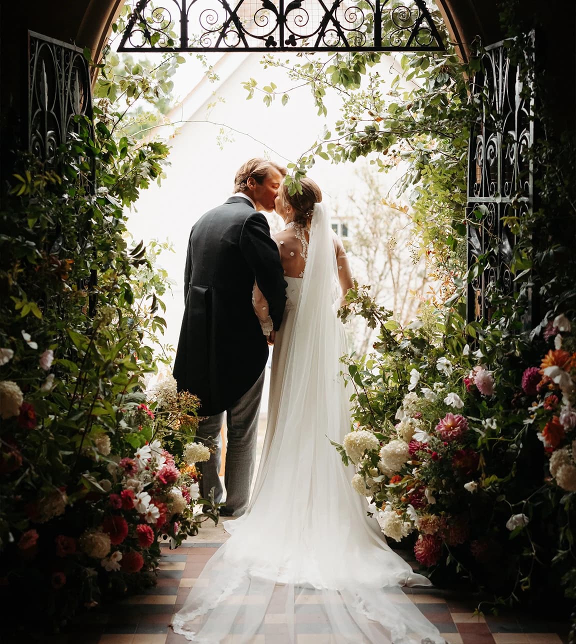 Newlyweds share a kiss in a church doorway framed by a floral wedding arch of dahlias and hydrangeas, her veil trailing behind
